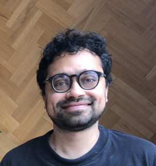 Headshot photo of speaker Dr. Siddique standing in front of a geometric pattern wood background. He is wearing a dark shirt, glasses, and smiling.