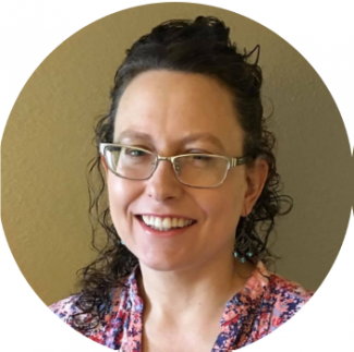 A photo of Dr. Jill Bradbury smiling. She has fair skin and curly hair, and is wearing glasses, dangly earrings, and a patterned top